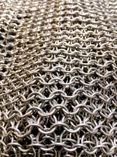 A close-up view of a tightly woven metal chainmail, showcasing the intricate interlocking rings that form a smooth, flexible surface. The pattern appears repetitive and dense, suggesting durability and strength.