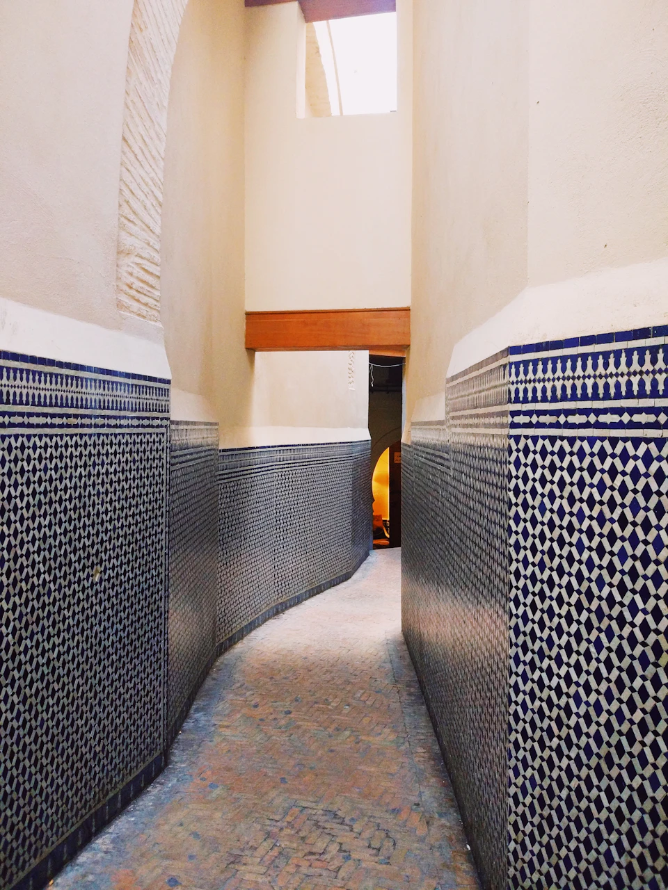 The image shows a narrow alleyway with blue and white tiled walls on both sides, creating an intricate pattern that adds visual interest to the scene. The floor is made of red bricks, providing a warm contrast to the cool tones of the tiles. On one side of the alleyway, there's a small archway leading into the distance, while on the other side, a window can be seen, allowing natural light to enter the space.

The perspective of the image is from ground level, looking down the length of the alleyway, giving it an immersive feel as if one could step right through and explore further. The precise arrangement of the tiled walls and floor creates a sense of depth in the scene, drawing the viewer's eye along the path.