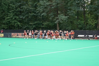 A group of athletes wearing uniforms are gathered on a green field with orange cones spread out in front of them. There is a black backdrop with the text 'FEAR THE' visible behind them and trees in the background. Some benches and equipment are also visible to the right side of the image.
