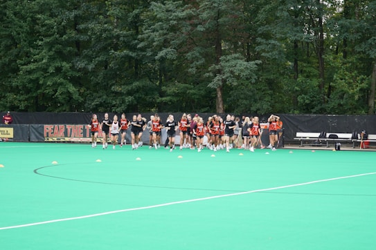 A group of athletes wearing uniforms are gathered on a green field with orange cones spread out in front of them. There is a black backdrop with the text 'FEAR THE' visible behind them and trees in the background. Some benches and equipment are also visible to the right side of the image.