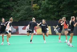 A group of young women in athletic gear are performing warm-up exercises on a green sports field, with trees in the background.