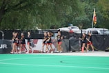 A group of athletes wearing black and red sports uniforms walk alongside a fence on a sports field. They are holding lacrosse sticks and are near a Maryland state flag. Trees and parked cars are visible in the background.