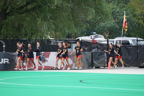 A group of athletes wearing black and red sports uniforms walk alongside a fence on a sports field. They are holding lacrosse sticks and are near a Maryland state flag. Trees and parked cars are visible in the background.