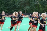 A group of young women wearing matching sports uniforms are jogging together on a green field. They are holding lacrosse sticks and appear to be part of a team, with 'Maryland' written on their shirts. The background is filled with lush green trees, suggesting an outdoor sports complex.
