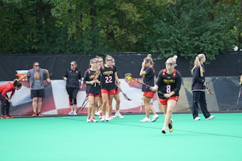 A group of female lacrosse players and coaches are on a field. The players are wearing black and red uniforms with the word 'Maryland' visible on their tops. Some of them hold lacrosse sticks as they walk and stand in various positions. In the background, there are trees and a fence with a flag-like design.