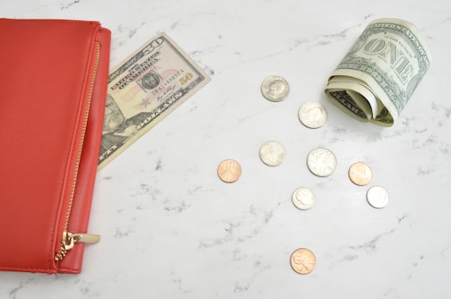 A red wallet with a golden zipper rests on a marble surface with a partially visible fifty-dollar bill sticking out. Nearby, a roll of one-dollar bills and loose change, including quarters and pennies, are scattered around.
