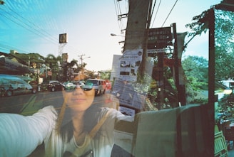 A double exposure photograph featuring a street scene with various cars and signs, overlaid with a woman's face reflected on a glass surface. The scene includes power lines, a street sign, and a vibrant, clear sky. The woman's face is partially obscured, and she appears to be wearing sunglasses.