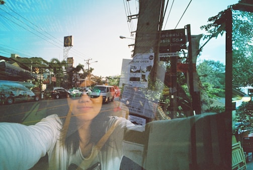 A double exposure photograph featuring a street scene with various cars and signs, overlaid with a woman's face reflected on a glass surface. The scene includes power lines, a street sign, and a vibrant, clear sky. The woman's face is partially obscured, and she appears to be wearing sunglasses.