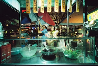 A street food stall with various ingredients displayed in glass containers, including limes and black jelly. Two people are working behind the counter, one holding a drink, while colorful signs hang overhead. The surrounding area is bustling with activity and illuminated by streetlights.
