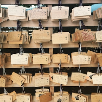 Wooden plaques with handwritten messages are displayed on a wooden rack, each hanging with a string. The plaques contain various writings and drawings in different styles and languages.