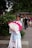A person is dressed in traditional attire, holding a large white and pink fan. The background shows a group of people walking and relaxing in an outdoor park setting, with lush greenery and trees surrounding them.