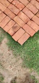 A neatly stacked arrangement of red bricks is placed on the ground, next to a patch of green grass and bare soil.