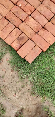A neatly stacked arrangement of red bricks is placed on the ground, next to a patch of green grass and bare soil.
