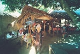 An outdoor market stall under a thatched roof, displaying colorful woven bags and textiles. A person in traditional attire stands in front of the stall. Sunlight filters through the leaves of nearby trees, creating dappled shadows on the ground.