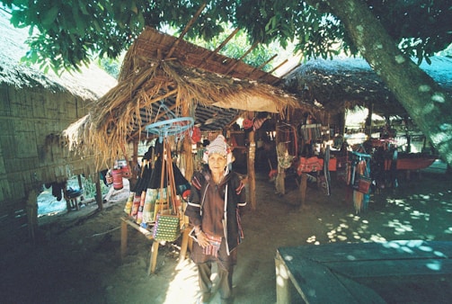 An outdoor market stall under a thatched roof, displaying colorful woven bags and textiles. A person in traditional attire stands in front of the stall. Sunlight filters through the leaves of nearby trees, creating dappled shadows on the ground.