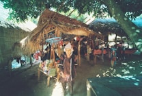 An outdoor market stall under a thatched roof, displaying colorful woven bags and textiles. A person in traditional attire stands in front of the stall. Sunlight filters through the leaves of nearby trees, creating dappled shadows on the ground.