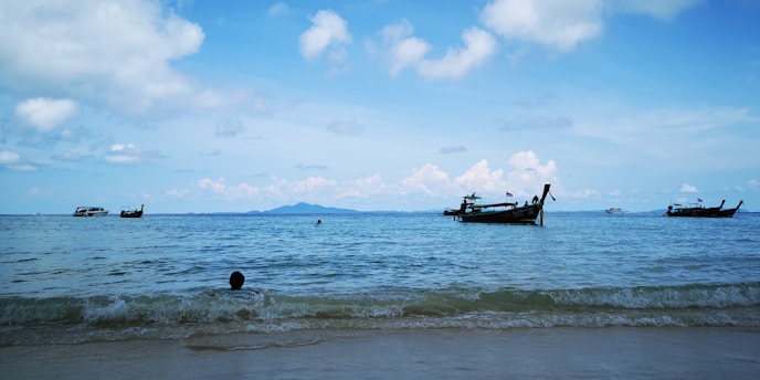 Several traditional long-tail boats float on a calm sea with a few swimmers near the shore against a backdrop of distant mountains and a partly cloudy blue sky.