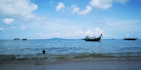 Several traditional long-tail boats float on a calm sea with a few swimmers near the shore against a backdrop of distant mountains and a partly cloudy blue sky.
