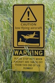 A yellow caution sign warns of low flying aircraft near a runway. It features an exclamation mark inside a triangle, an image of an aircraft, and instructions for stopping when aircraft are taking off.