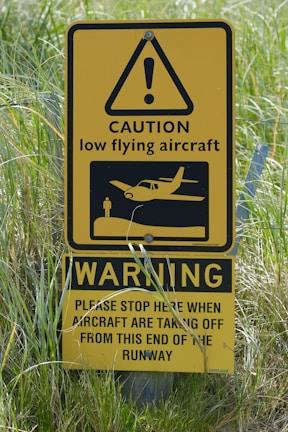 A yellow caution sign warns of low flying aircraft near a runway. It features an exclamation mark inside a triangle, an image of an aircraft, and instructions for stopping when aircraft are taking off.