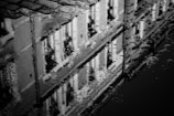 A black and white reflection of a building in a puddle, featuring cobblestones and ripples on the water's surface. Texture is highlighted by the raindrops creating patterns in the water.