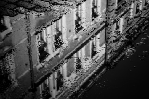 A black and white reflection of a building in a puddle, featuring cobblestones and ripples on the water's surface. Texture is highlighted by the raindrops creating patterns in the water.