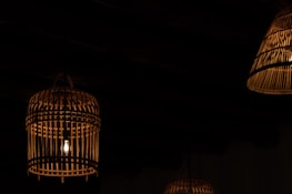 Dimly lit environment featuring several hanging lanterns made of wicker or bamboo. A soft glow radiates from the bulbs enclosed within these ornate, cage-like structures, casting subtle shadows against a dark background.