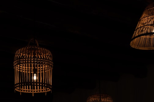 Dimly lit environment featuring several hanging lanterns made of wicker or bamboo. A soft glow radiates from the bulbs enclosed within these ornate, cage-like structures, casting subtle shadows against a dark background.