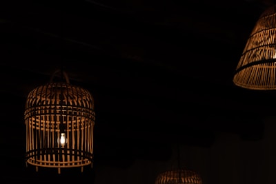 Dimly lit environment featuring several hanging lanterns made of wicker or bamboo. A soft glow radiates from the bulbs enclosed within these ornate, cage-like structures, casting subtle shadows against a dark background.
