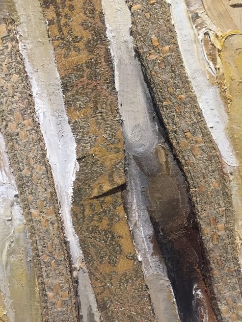 The image features a close-up view of textured surfaces with alternating strips of fabric and paint. The fabric strips have intricate patterns resembling floral and geometric designs in earthy tones. The painted areas seem layered, showing brush strokes in beige, white, and brown shades, creating a tactile and rich surface.