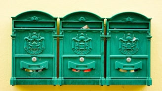 Three green, ornate mailboxes are mounted on a pale yellow wall. Each mailbox features an intricate coat of arms design and keyholes. Pieces of mail can be seen peeking out from the slots.