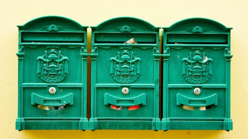 Three green, ornate mailboxes are mounted on a pale yellow wall. Each mailbox features an intricate coat of arms design and keyholes. Pieces of mail can be seen peeking out from the slots.