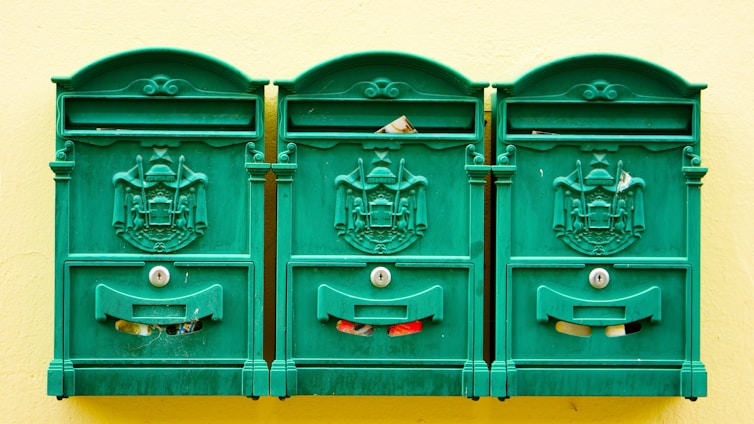 Three green, ornate mailboxes are mounted on a pale yellow wall. Each mailbox features an intricate coat of arms design and keyholes. Pieces of mail can be seen peeking out from the slots.