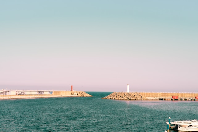 A serene harbor scene featuring calm blue waters meeting a clear sky with a gentle gradient of pastel pink and blue. Two breakwaters extend into the water, each topped with a lighthouse&mdash;one white and one red. In the foreground, part of a docked boat is visible.