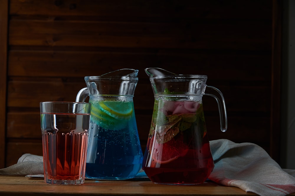 two clear glass pitchers with red and blue liquid on table beside glass filled with red liquid