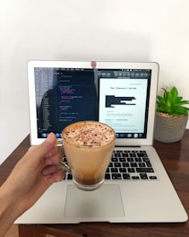 A hand holds a glass mug filled with a frothy coffee, sprinkled with chocolate, in front of an open laptop displaying code on one side and a document titled 'The Fibonacci Series' on the other. A small green potted plant is placed on the right side of the laptop on the wooden table.