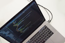 A laptop displaying a text editor with programming code on the screen. The keyboard is visible, along with a connecting cable, all against a white background.