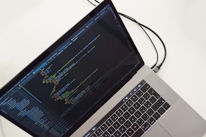 A laptop displaying a text editor with programming code on the screen. The keyboard is visible, along with a connecting cable, all against a white background.