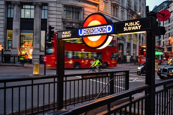 A busy city street scene features an entrance to a subway station with the iconic Underground sign. Double-decker red buses and taxis move along the road, while a cyclist dressed in a bright green jacket rides past. Tall buildings line the street, and the lighting suggests early evening.