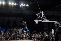 A basketball player is in mid-air performing a slam dunk in a crowded indoor arena. The player is holding a basketball and is about to score in the hoop. The crowd in the background watches intently, with some spectators capturing the moment with their phones.