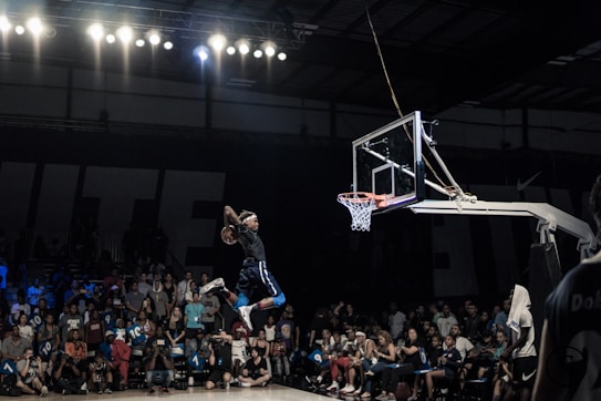 A basketball player is in mid-air performing a slam dunk in a crowded indoor arena. The player is holding a basketball and is about to score in the hoop. The crowd in the background watches intently, with some spectators capturing the moment with their phones.