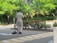A person wearing a light-colored uniform is sweeping the ground with a broom and dustpan in an outdoor area bordered by green bushes and red flowers. Tall trees with lush green foliage form the background, creating a serene and natural atmosphere.