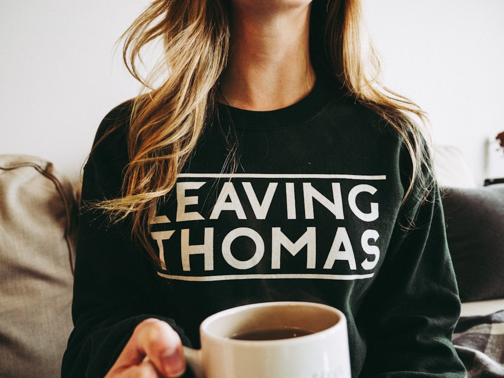 woman wearing black Leaving Thomas-printed sweater while holding white ceramic mug filled with coffee