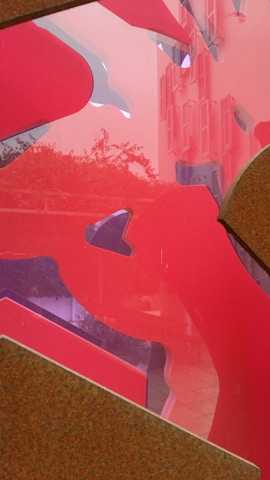 Abstract shapes in shades of red overlay the image, with a hint of a building and foliage visible in the background through a translucent section. The combination of sharp edges and flowing forms creates a dynamic visual experience.