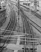 A black and white photograph of multiple railway tracks intersecting and diverging, surrounded by electrical wires and poles. The tracks extend into the distance flanked by gravel and a stone wall on one side.