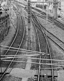 A black and white photograph of multiple railway tracks intersecting and diverging, surrounded by electrical wires and poles. The tracks extend into the distance flanked by gravel and a stone wall on one side.