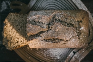 A loaf of bread with a rustic, homemade appearance is placed on a wooden board. The bread has a crusty texture with visible cracks and is dusted with flour. A slice has been cut from the loaf, revealing the soft, crumbly interior.