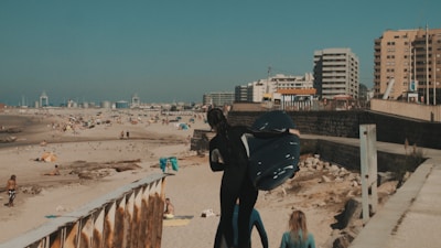 A busy beach scene with numerous people scattered across the sandy shore. The focal point is a person in a wetsuit holding a surfboard, walking down a pathway alongside another individual. The background features tall buildings and city infrastructure, extending towards the horizon.
