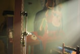 A stage stand is positioned prominently in the foreground with light shining through, casting soft illumination. In the background, there is a mural depicting abstract figures, possibly a religious or ceremonial scene. A fire extinguisher is mounted on a wall to the left, and part of a chair is visible at the bottom right.
