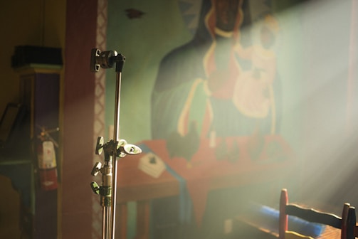 A stage stand is positioned prominently in the foreground with light shining through, casting soft illumination. In the background, there is a mural depicting abstract figures, possibly a religious or ceremonial scene. A fire extinguisher is mounted on a wall to the left, and part of a chair is visible at the bottom right.